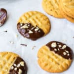 Close up of 3 Dark Chocolate Dipped Peanut Butter Cookies with chopped peanuts on white parchment paper with a few undipped cookies and a cooling rack in the background