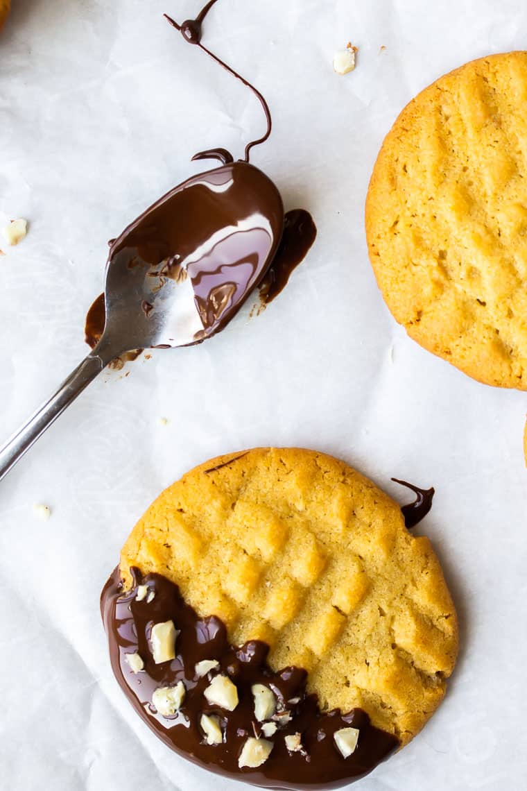 Close up of a chocolate dipped peanut butter cookie covered in chopped peanuts with a second cookie and a chocolate covered spoon in the background