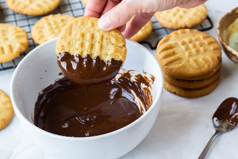 A hand dipping a peanut butter cookie into melted chocolate with a spoon and cooling rack full of more cookies nearby