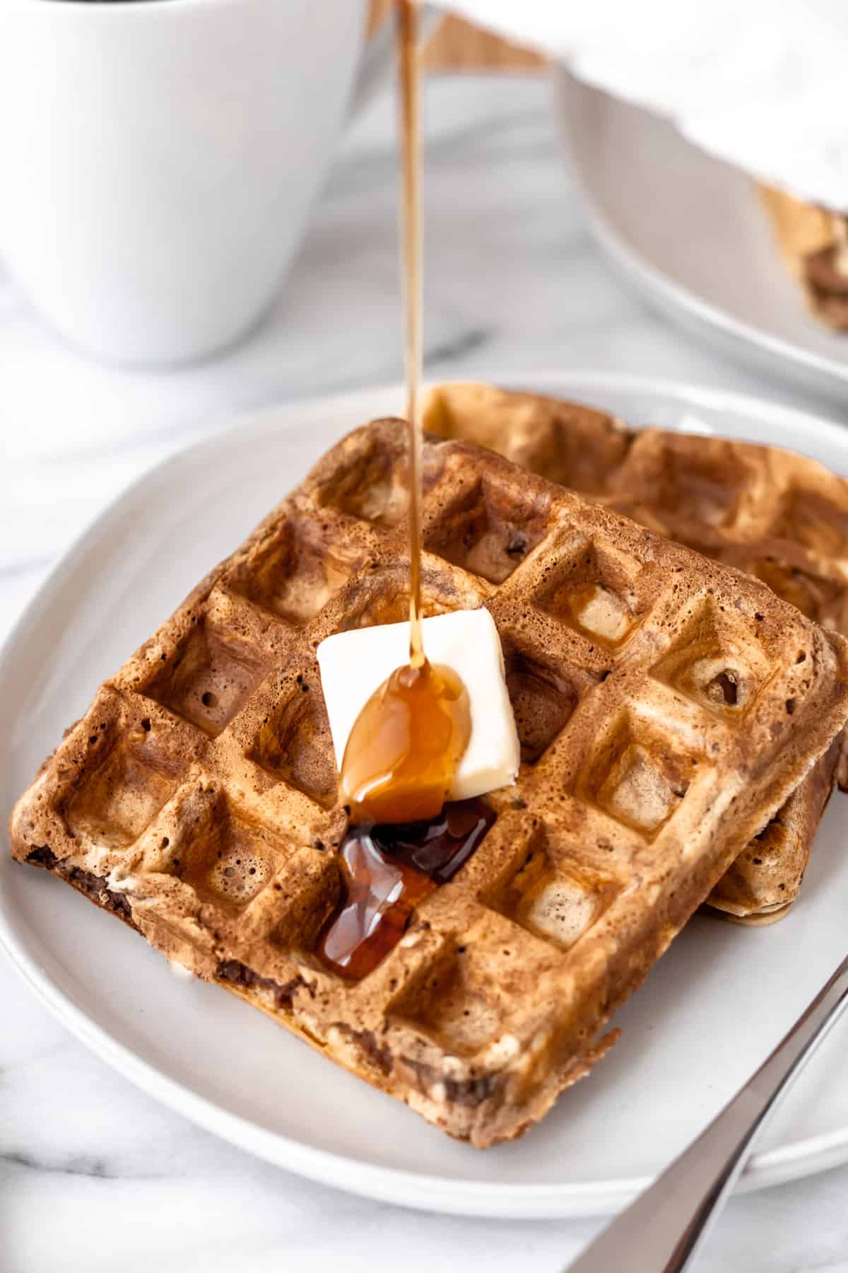 Maple syrup being poured onto chocolate peanut butter waffles on a white plate.