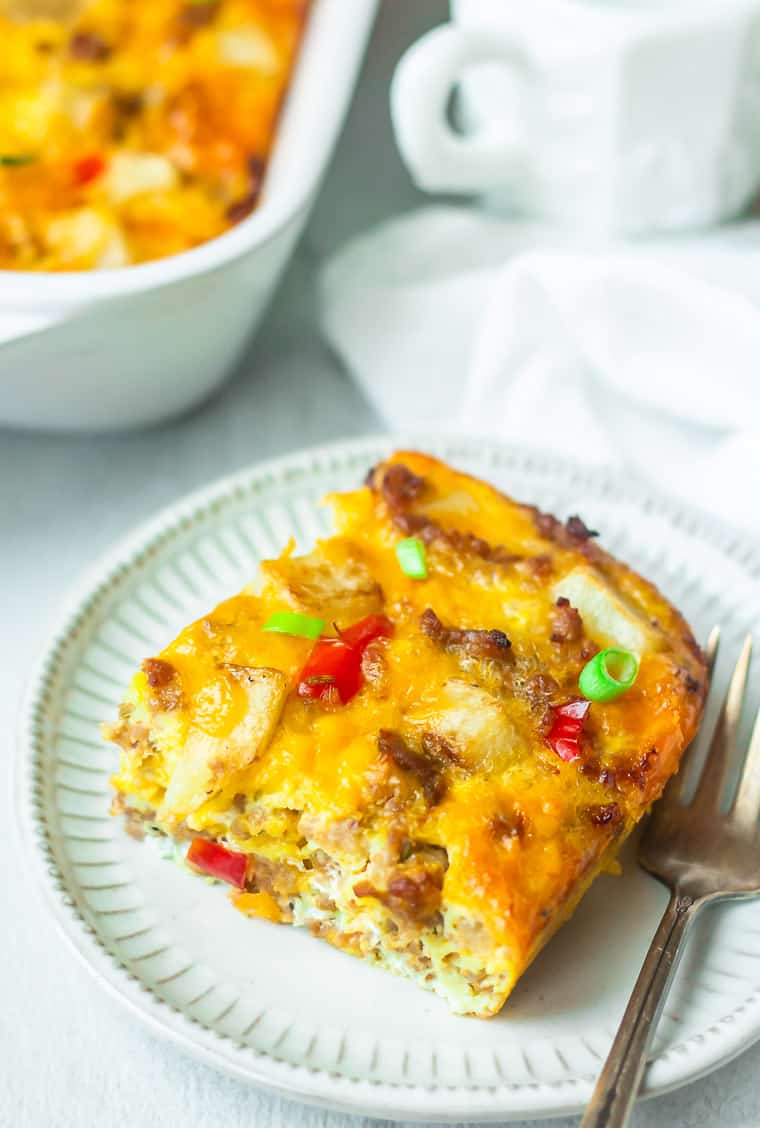 A slice of Sausage Breakfast Casserole on a plate with a fork with part of the casserole dish, a white cup, and a white napkin in the background