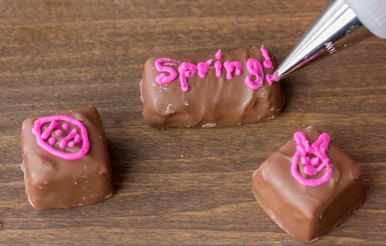 Piping pink butterncream designs and words onto pieces of chocolate candy