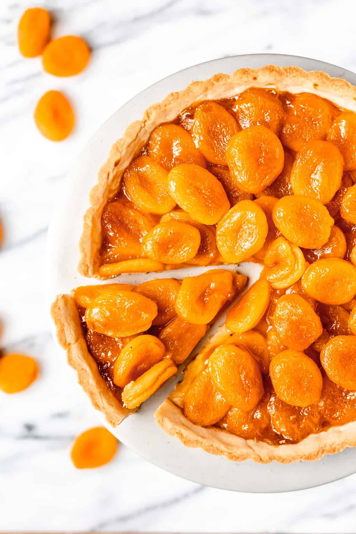 Overhead view of an apricot tart with one slice pulled away a little and apricots on the table around it.