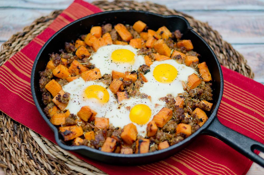 Sweet Potato Hash with Sausage and 4 eggs in a black skillet on a red towel on a wicker mat