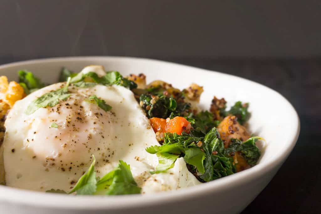 Quinoa Breakfast Hash Topped with an Egg in a White Bowl with smoke coming off the top - all on a gray background
