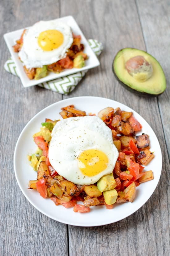 Avocado Bacon Hash with Eggs on a white plate with a second smaller serving in the background and an avocado half - on a gray wood background