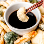 Close up of a piece of vegetable tempura being held up with chopsticks over a bowl of sauce.
