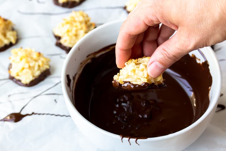 A hand dipping a coconut macaroon into a white bowl of chocolate with more macaroons and drizzled chocolate spills in the background