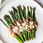 Overhead of prosciutto wrapped asparagus topped with Parmesan cheese and almonds on a white plate over a white background with a white napkin