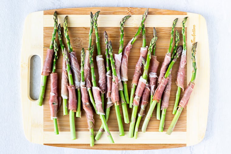 Prosciutto wrapped asparagus on a wood cutting board over a white background