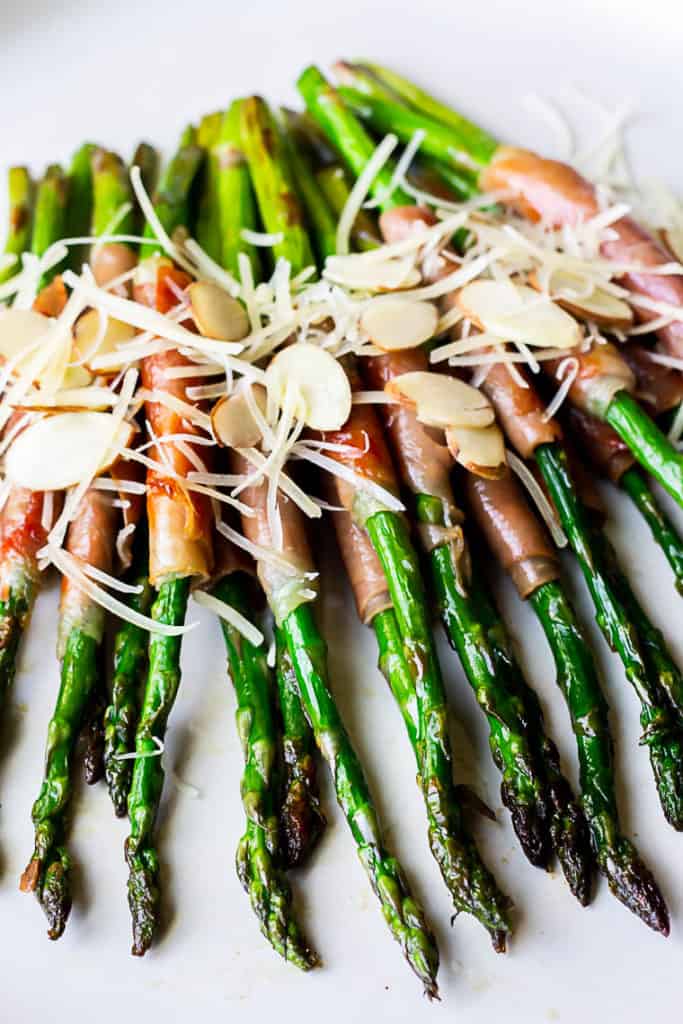 Close up of asparagus wrapped in prosciutto and topped with sliced almonds and shredded parmesan cheese on a white plate