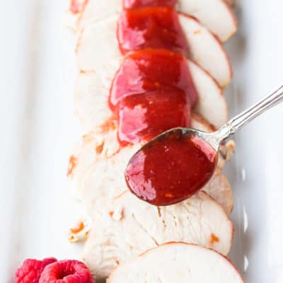 Spooning raspberry barbecue sauce over sliced meat on a white tray with raspberries on the side