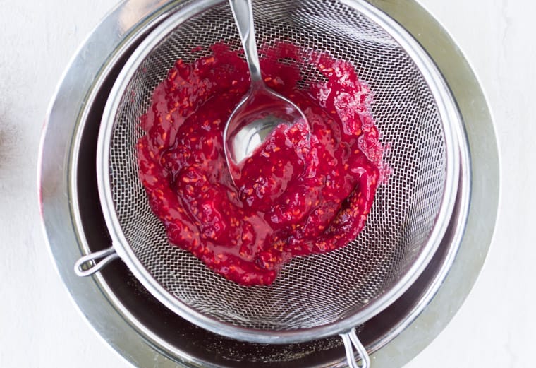 Pressing raspberries through a mesh strainer with a spoon over a white background