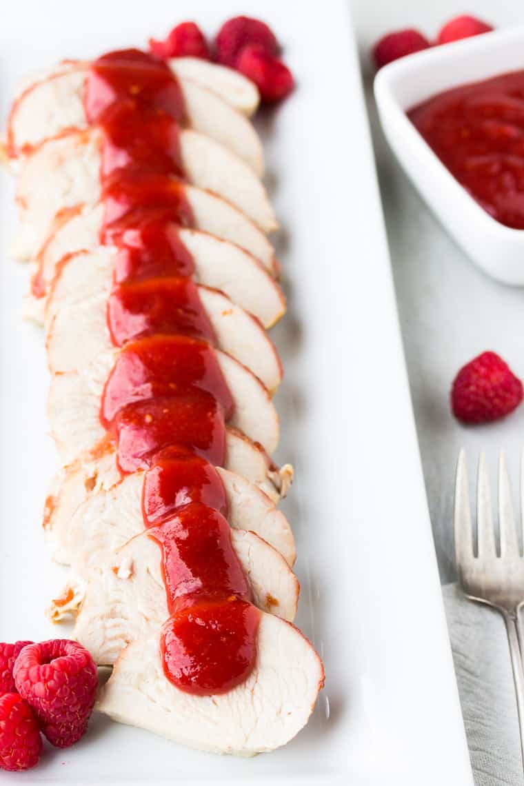 A white tray of sliced meat with raspberry barbecue sauce over it and in a small bowl off to the side with extra raspberries and a small fork in the background