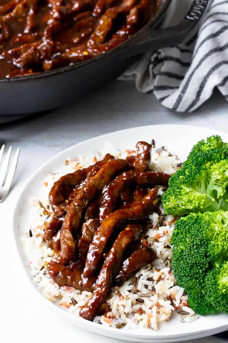A white palte with rice, mongolian beef, and broccoli with a cast iron pan of beef and a black and white napkin in the background