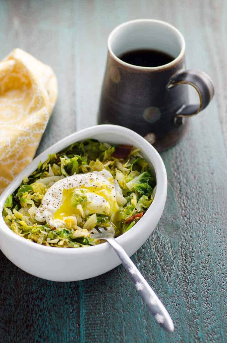 Shredded Brussels topped with an egg in a white bowl with a fork on a gray background with a cup of coffee in a brown mug and a yellow napkin off to the side