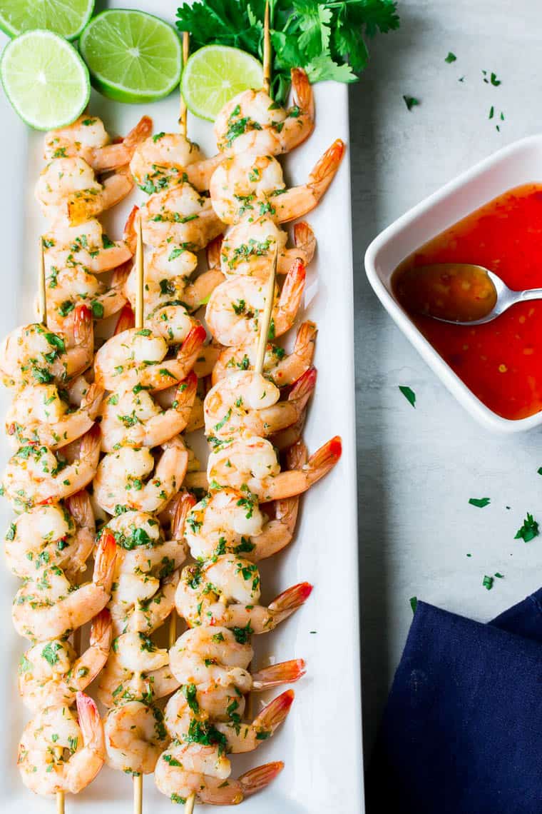 Sweet Chili Lime Shrimp Skewers on a white tray with cilantro and lime, a small dish of extra sauce, and a blue napkin
