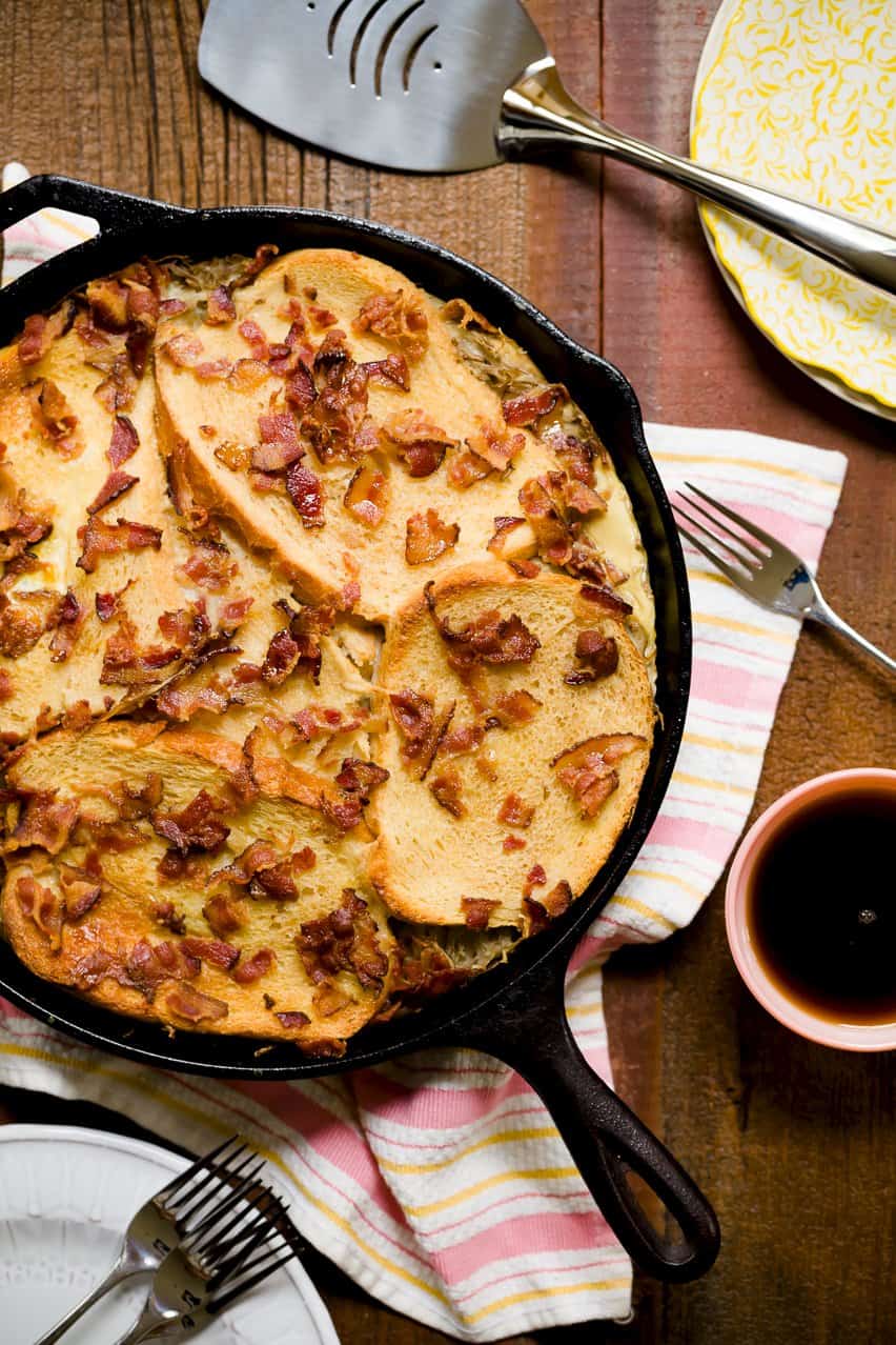 French Toast in a Black Skillet with pieces of bacon on top. All over a wood table with a spatula, plates, forks, a pink and white towel and a cup of syrup