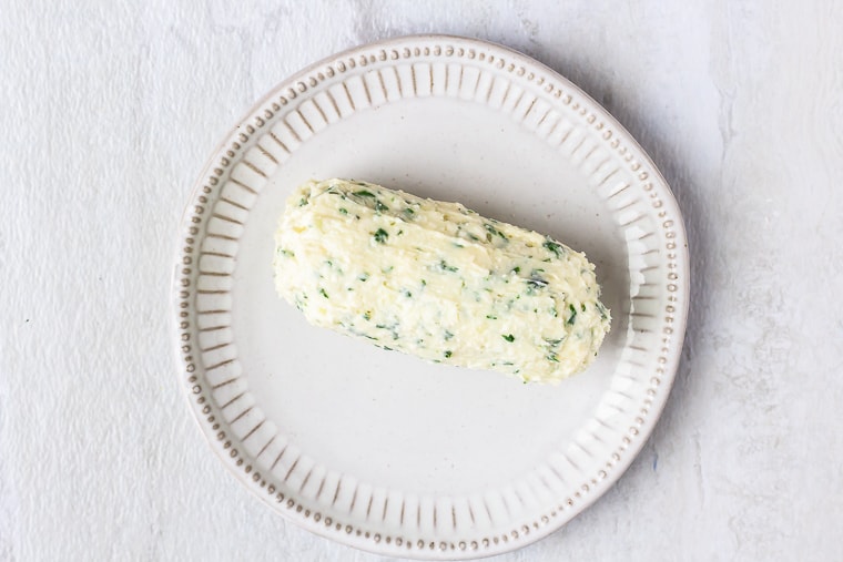 A log of herb butter on a white plate over a white background