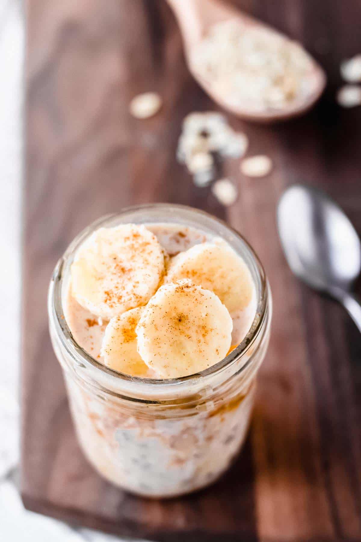 angles view of a mason jar of peanut butter and banana overnight oats on a wood board with wood spoon and a spoon blurred in the background