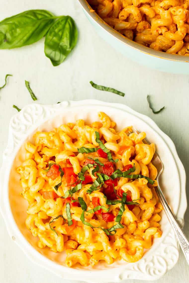 A white bowl filled with creamy roasted red pepper pasta and topped with diced peppers and fresh basil with basil leaves in the background and a pot of more pasta over a white background