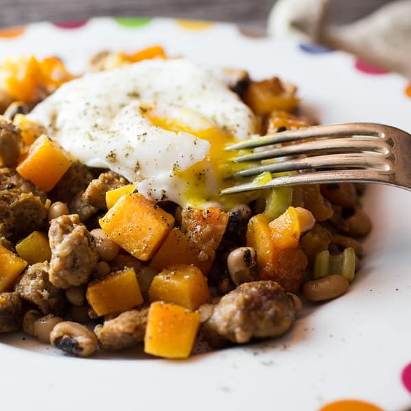 Close up of butternut squash and sausage on a white plate topped with an egg being broken open with a fork