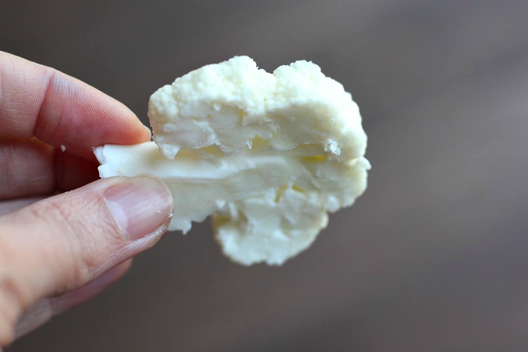 A hand holding out single piece of Cauliflower Sliced Thin over a gray background