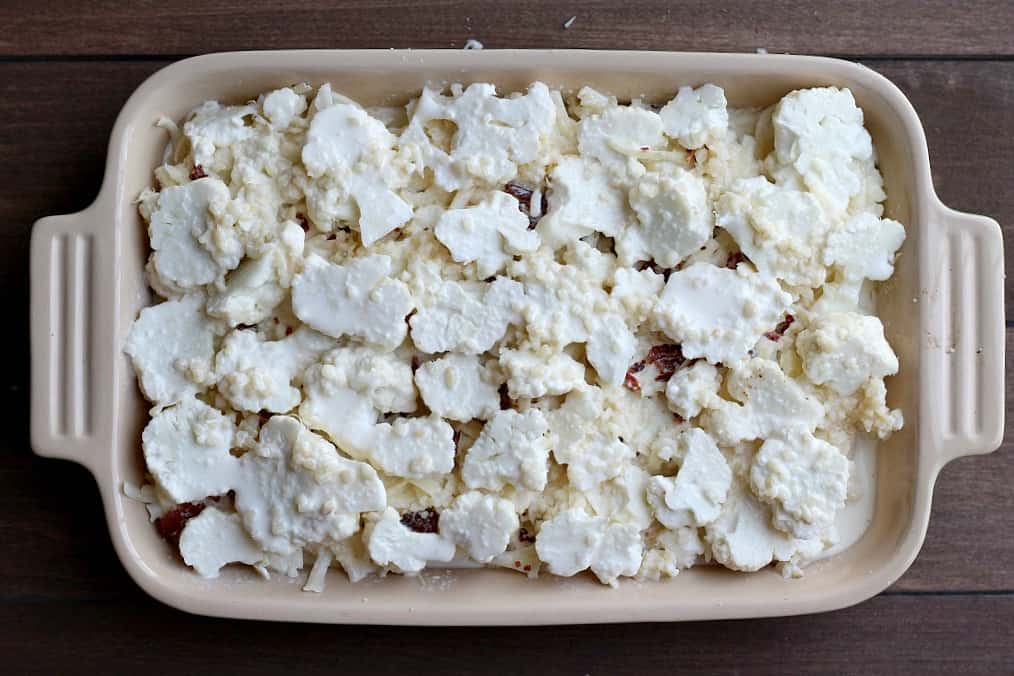 Cauliflower with layers of cheese and bacon between it and cream sauce around it in a casserole dish over a wood background