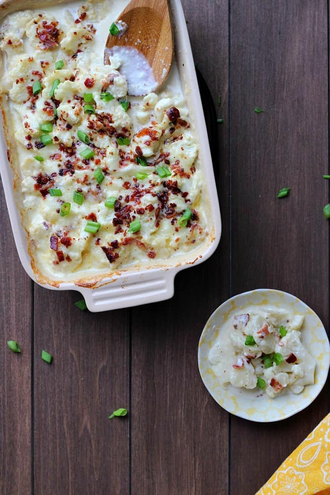 Baked keto Au Gratin Cauliflower in a casserole dish with a wood spoon and a smaller plate with a single serving on it all on a wood table with a few green onions scattered around