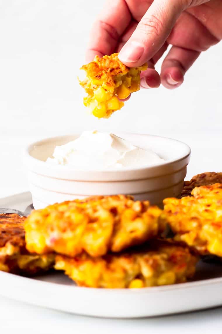A piece of corn fritter being held up over a bowl of sour cream on a plate with more corn fritters