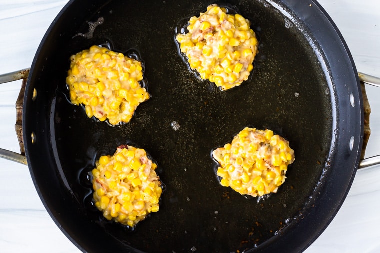 4 corn fritters cooking in oil in a black skillet over a white background