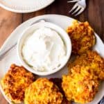 Corn Fritters on a white plate with a white bowl of sour cream, on a wood board with a small white plate with a single fritter on it and 2 forks next to it