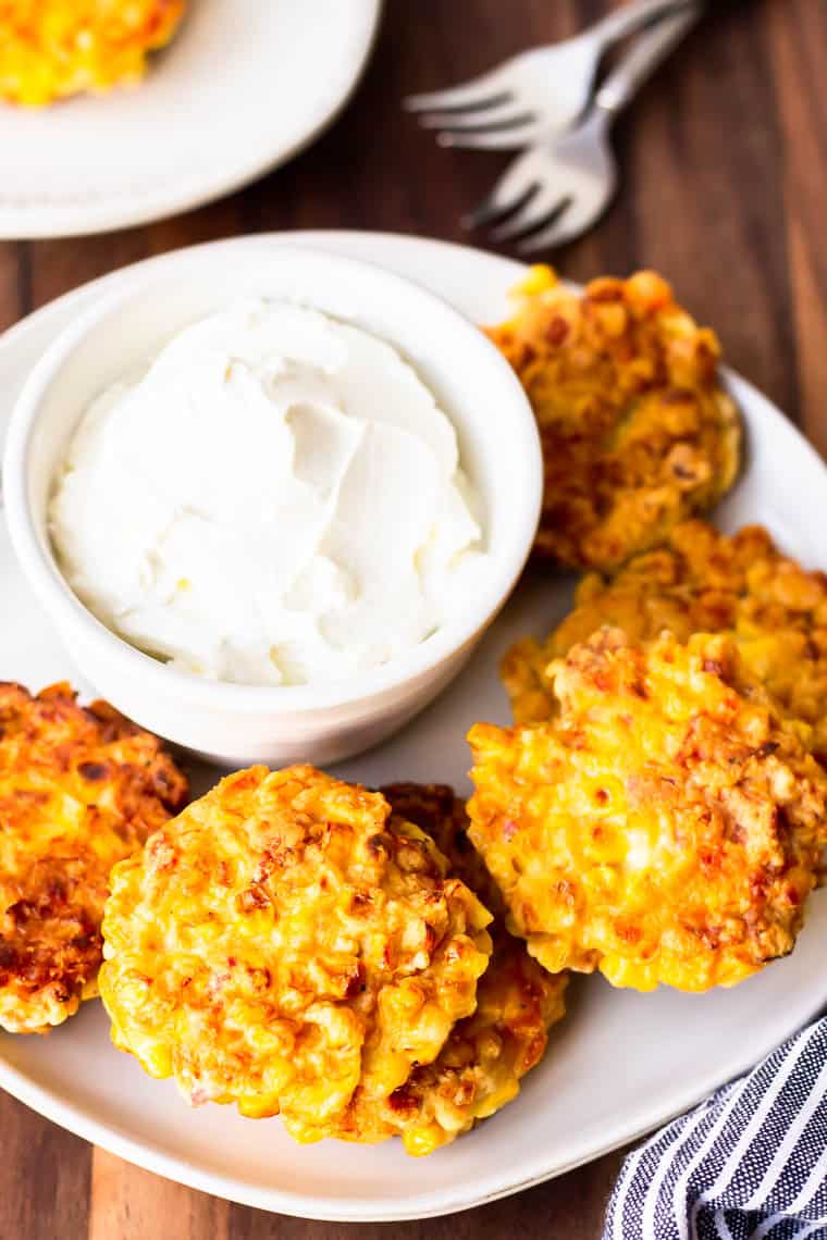 A plate of fried corn cakes with a bowl of sour cream on a wood board with a second plate and 2 forks behind it