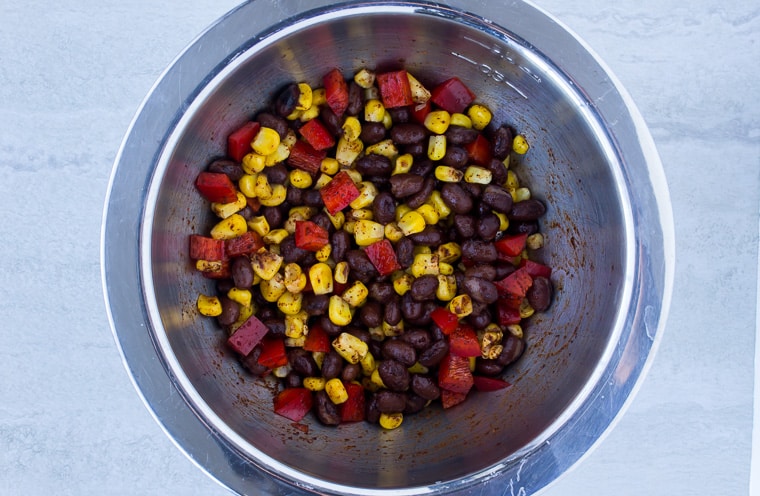 Corn, black beans, and diced red pepper mixed with chili powder in a small silver bowl.