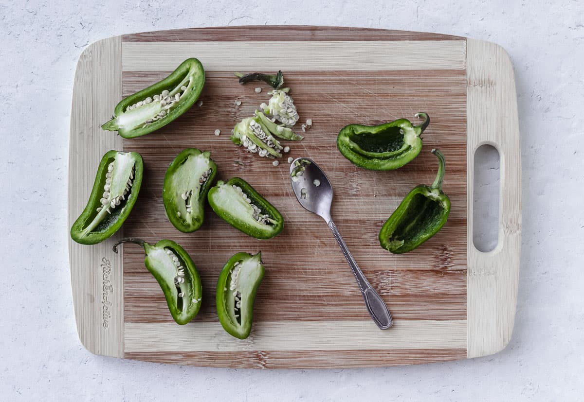 Jalapeno peppers on a wood board cut in half with the seeds removed from some and a spoon with them