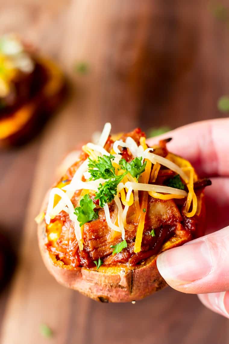 Holding up a single sweet potato round topped with pulled pork, cheese, and herbs with a wood background