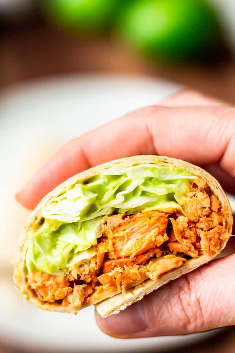 A hand holding up a Blackened Salmon Wrap filled with blackened salmon and avocado slaw with tortillas, limes and a wood table blurred in the background