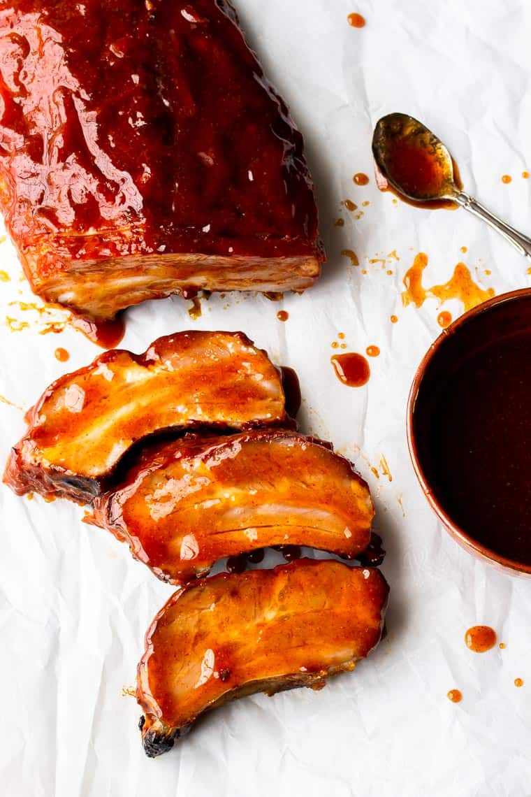 Chipotle Honey-Barbecue Ribs with 3 slices cut off, a dark bowl of barbecue sauce and a spoon on a white background with extra sauce spilled