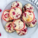 A white circle plate stacked with raspberry muffins with 4 loose raspberries and a white and cream kitchen towel in the background all on a white backdrop