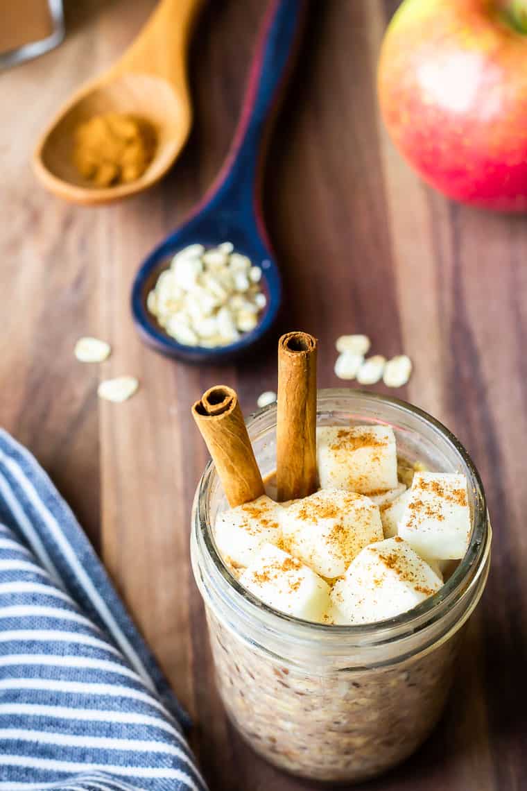 A mason jar of Apple Pie Overnight Oats with chopped apples and cinnamon sticks in it and a blue and white napkin, blue spoon with oats, brown spoon with cinnamon, and an apple in the background on a wood backdrop