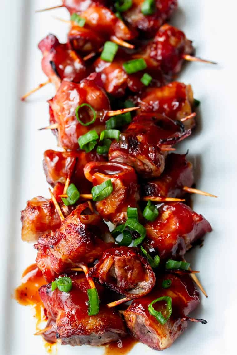 A white serving tray full of bacon wrapped barbecue beef bites topped with green onions