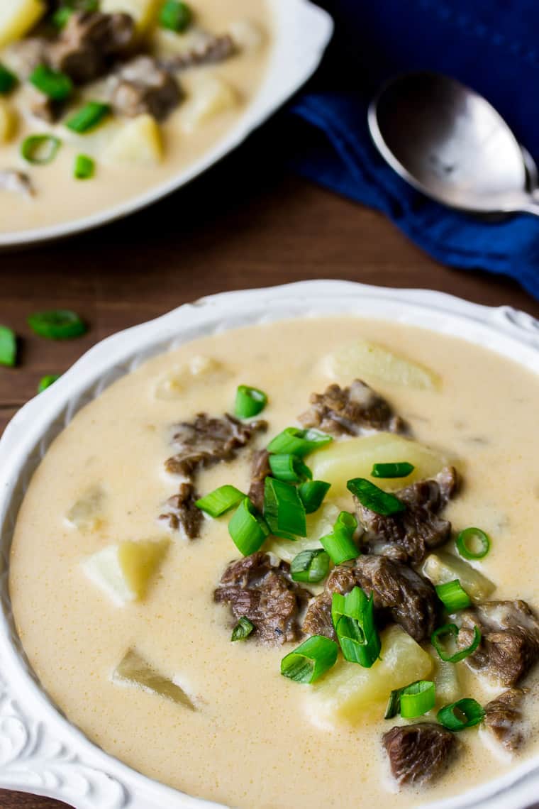 Close up of a bowl of Creamy Steak and Potato Soup topped with green onions with a second bowl in the background, a blue napkin and a spoon all on a wood background