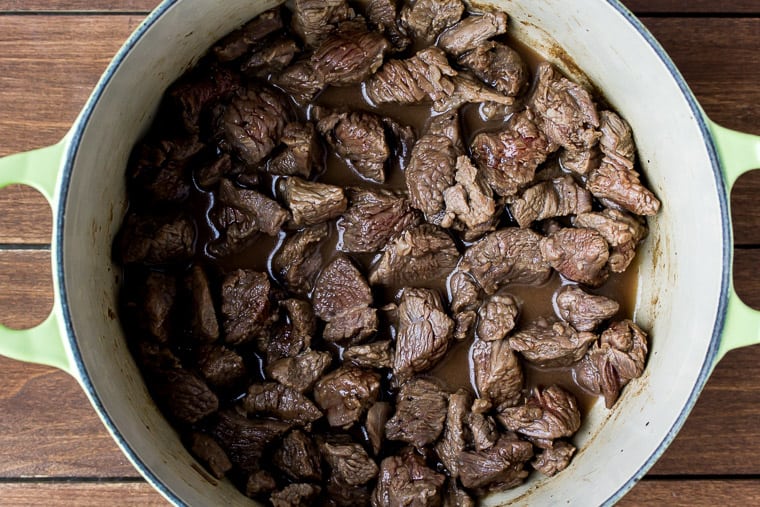 Beef cubes cooking in a cream color Dutch oven on a wood background