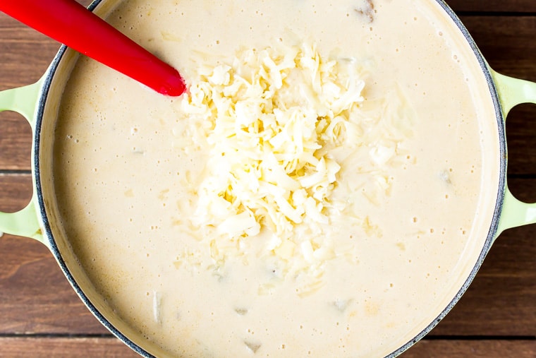 Shredded cheese added to a cream color Dutch oven with a cream based soup in it on a wood background