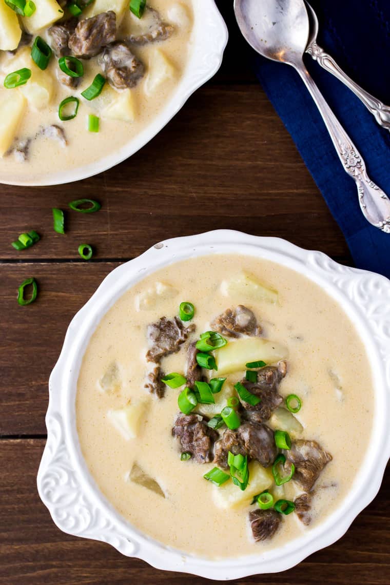 An overhead view of two bowls of Creamy Steak and Potato Soup topped with slices of green onion with a dark blue napkin and 2 spoons on a wood background