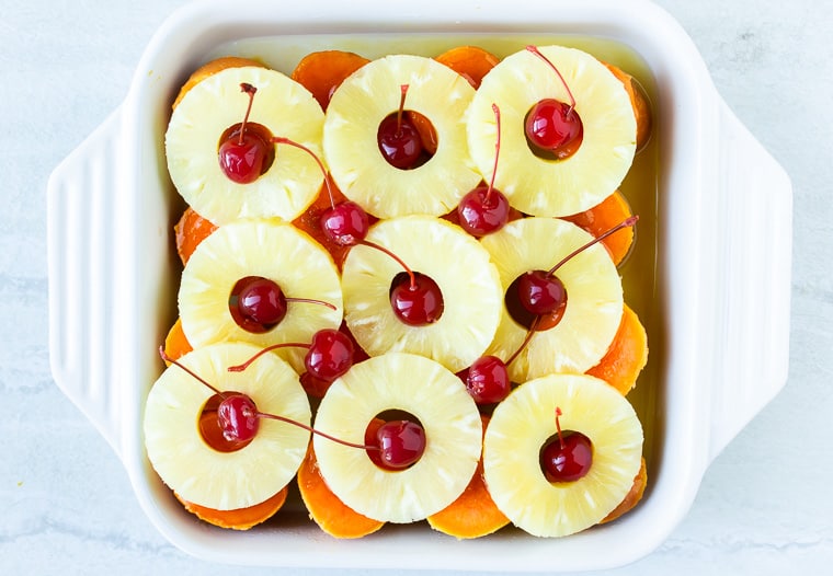 Slices of sweet potatoes in a white, square casserole dish and topped with pineapple rings and maraschino cherries