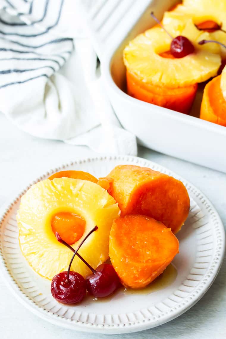 Candied Sweet Potatoes with Pineapple Slices and Cherries on a small white plate and in a white casserole dish in the background with a black and white striped napkin