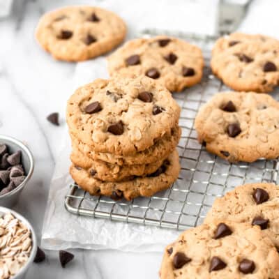 A stack of peanut butter chocolate chip oatmeal cookies on a cooling rack with more cookies, a mug and a white towel around it.