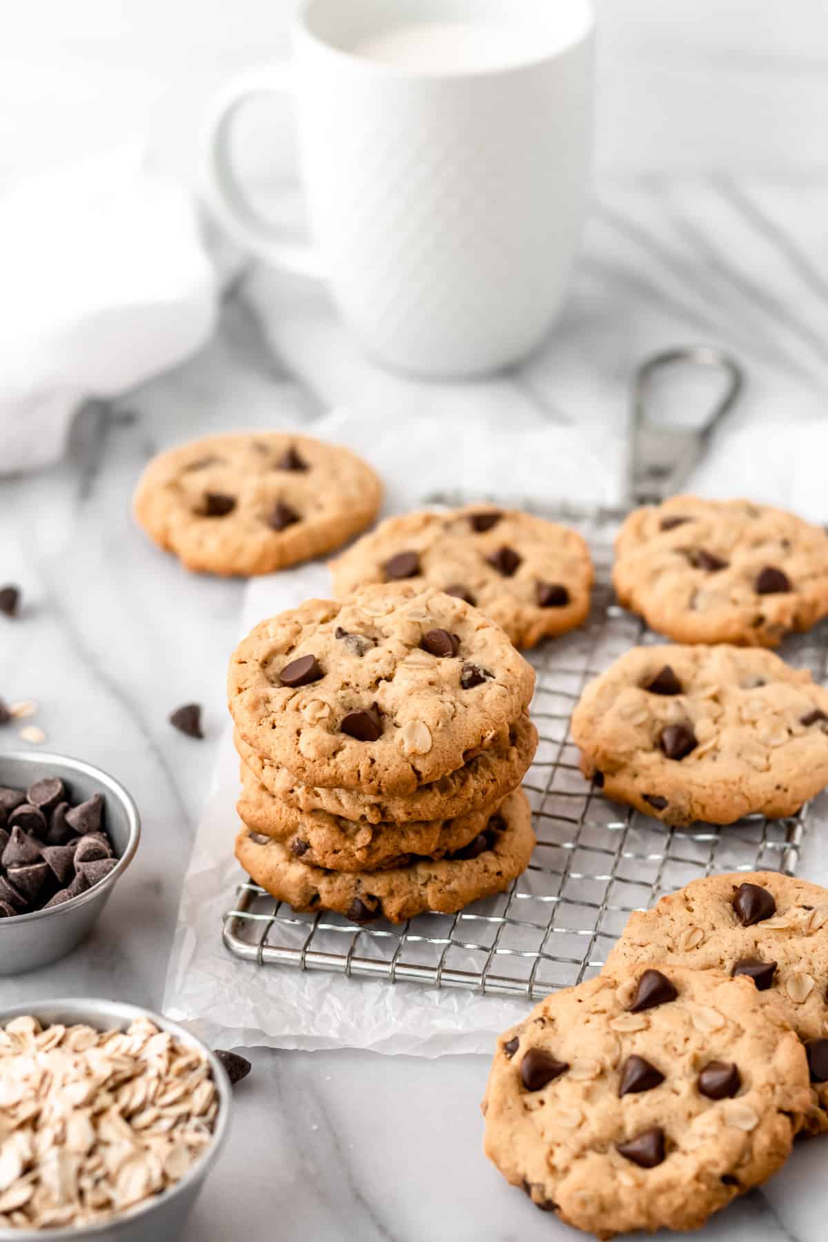 A stack of peanut butter chocolate chip oatmeal cookies on a cooling rack with more cookies, a mug and a white towel around it.