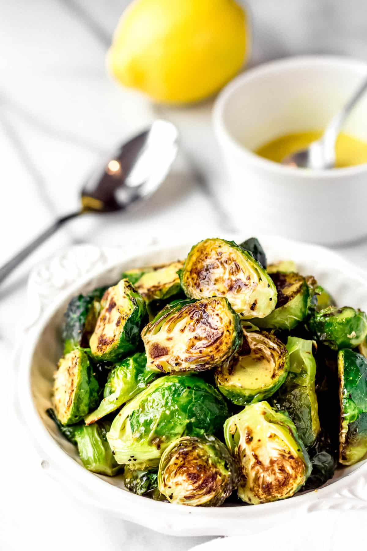 Roasted brussels sprouts in a white bowl with a lemon, spoon and small bowl of dressing in the background